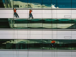 Professional window cleaners in safety harnesses cleaning the exterior windows of a modern glass office building. The workers are wearing orange safety shirts and helmets while suspended on ropes, with reflections of the cityscape and architectural details visible in the glass panels.